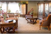 A bright, traditionally furnished sitting room with upholstered sofas and chairs, a round coffee table, floral arrangements, and a glass-front display cabinet by a window.
