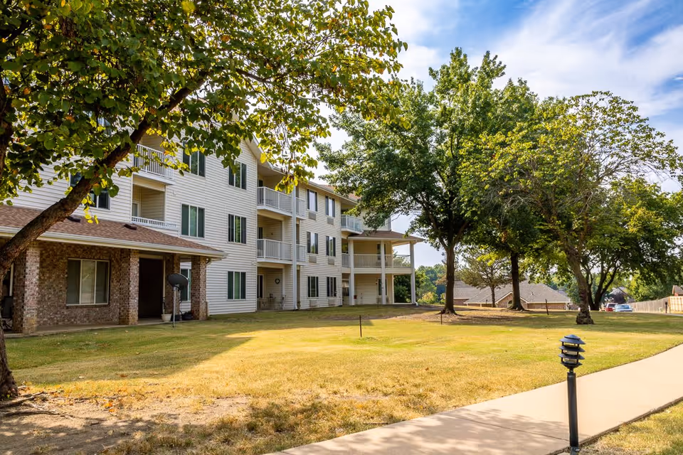 Exterior view of a three-story residential building with white siding and brick accents, surrounded by green trees and a well-maintained lawn. A paved walkway with a small lamp post runs alongside the building under a partly cloudy sky.