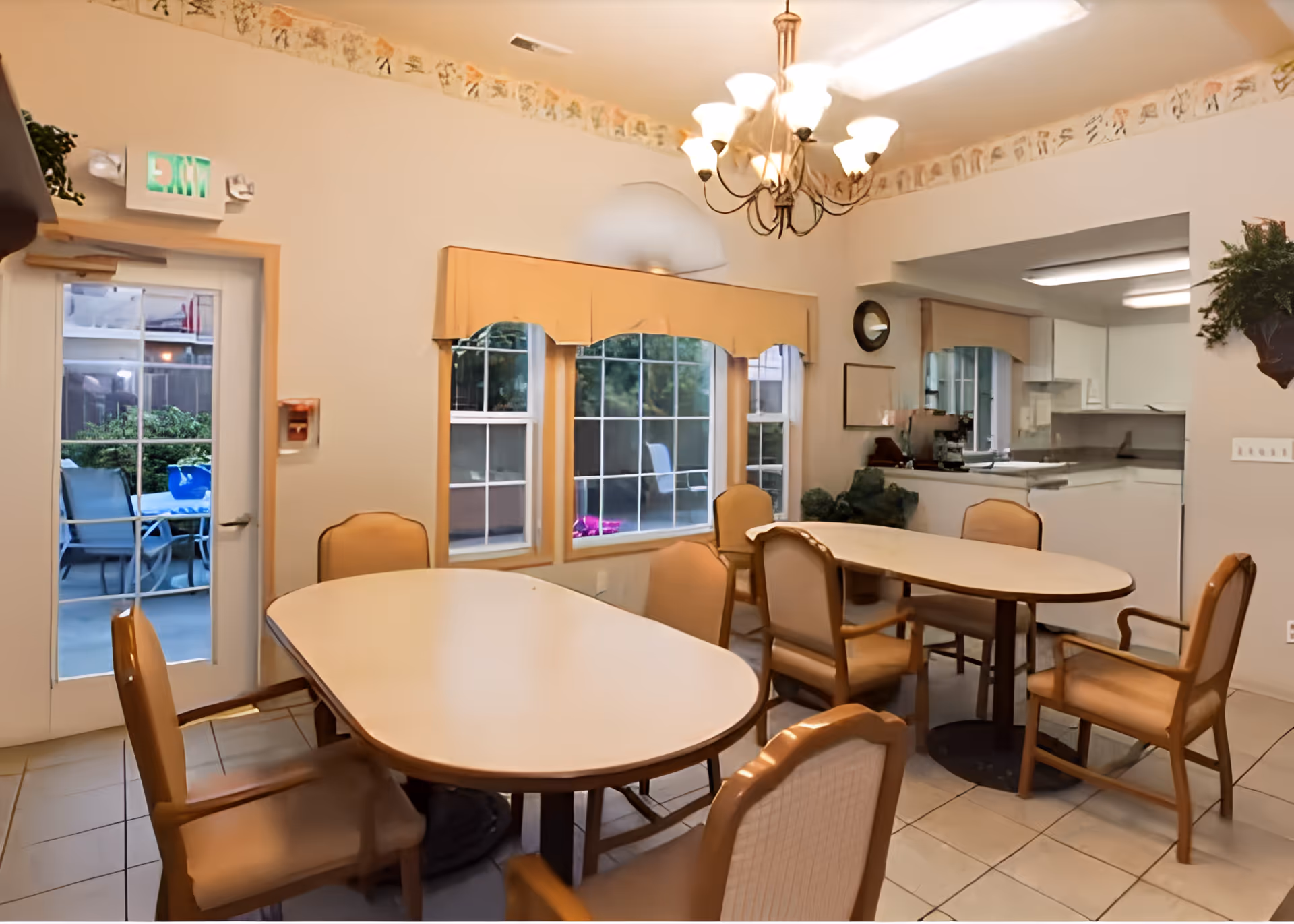 A dining area inside a senior living facility with two oval tables surrounded by cushioned chairs. There is a chandelier hanging from the ceiling, windows with valances, and a door leading to an outdoor patio with additional seating. The room has tiled floors and a partial view of a kitchen area in the background.
