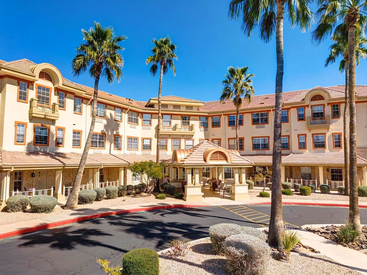 Exterior view of a three-story assisted living facility with beige walls and red-tiled roof, surrounded by palm trees and desert landscaping under a clear blue sky.
