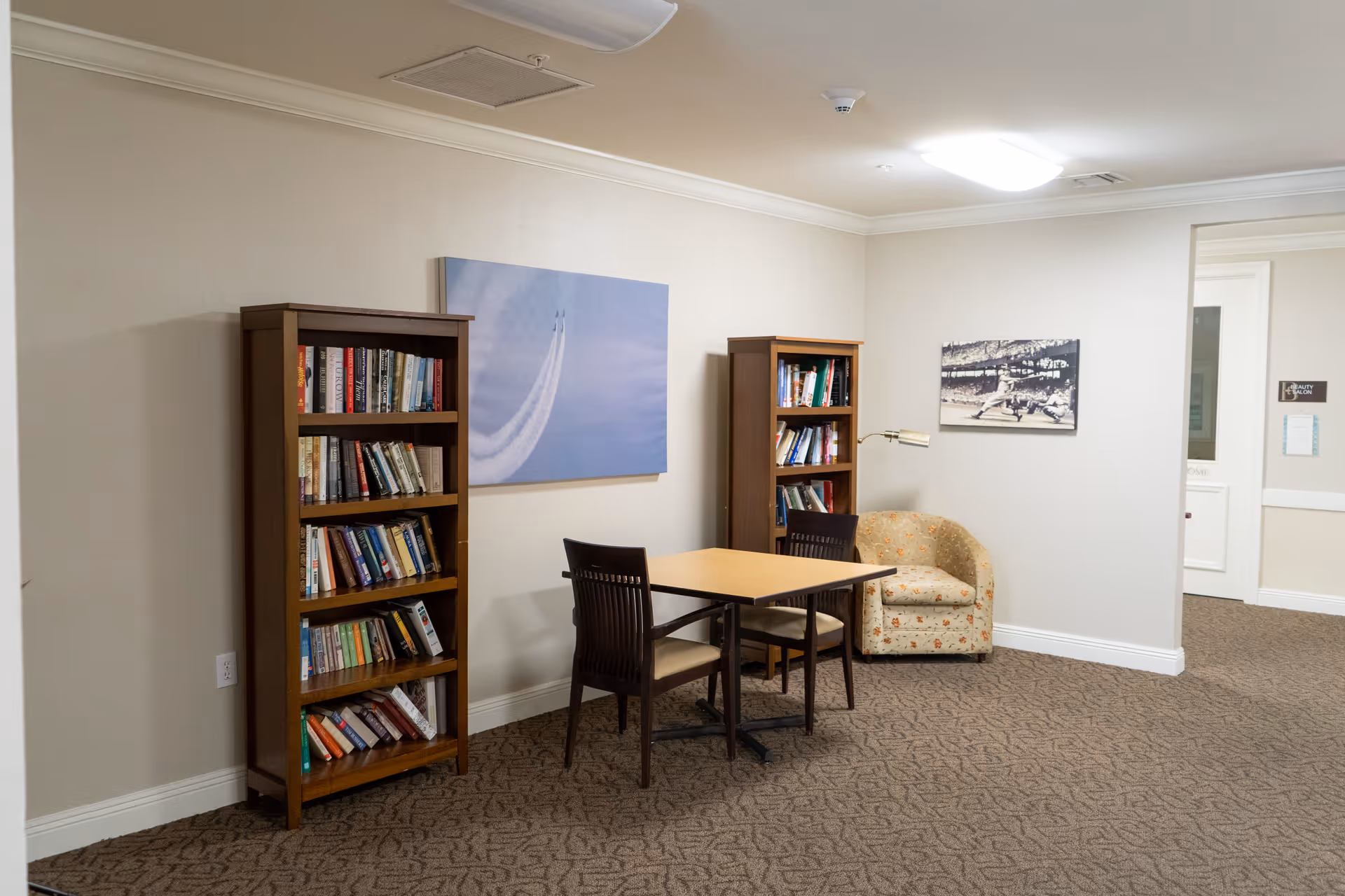 A quiet reading and seating area in a senior living facility with two wooden bookshelves filled with books, a small table with two chairs, and a floral-patterned armchair. The walls are decorated with two pictures, one showing airplanes flying in formation and the other a vintage baseball scene. The carpet is patterned, and there is a door labeled 'Beauty Salon' in the background.