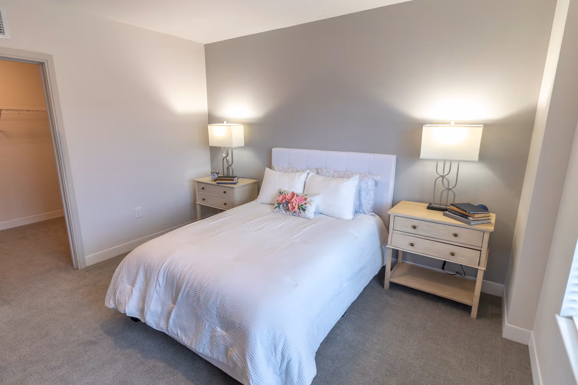 A neatly made bed with white bedding and decorative pillows in a bedroom. There are two wooden nightstands on either side of the bed, each with a lamp and some books. The walls are painted light gray and beige, and there is an open doorway leading to a closet with a wire shelf. The room has carpeted flooring and a window letting in natural light.