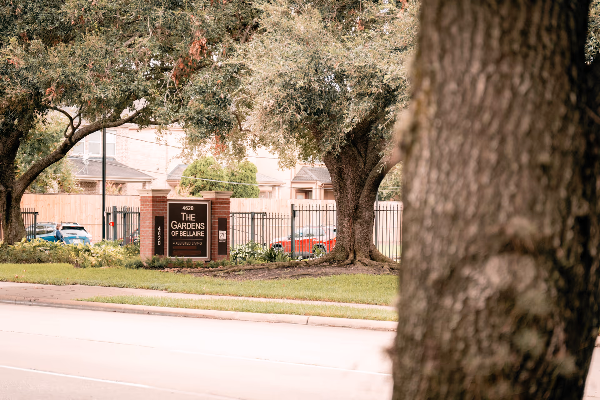 View of the entrance sign for The Gardens of Bellaire assisted living facility, surrounded by trees and greenery, with a street in the foreground and residential houses in the background.
