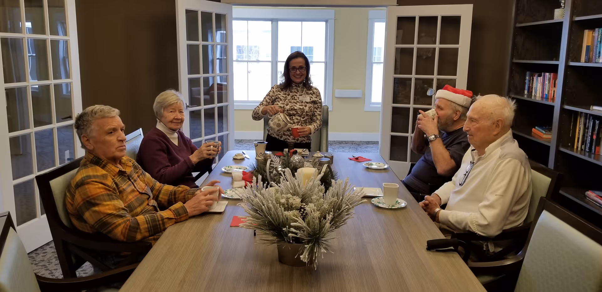 Five elderly people sitting around a wooden table in a well-lit room with large windows and glass-paneled doors. One woman is standing and pouring a beverage into a cup, while the others are seated, holding cups and engaging in conversation. The table is decorated with a frosted floral centerpiece and holiday-themed napkins. One man is wearing a Santa hat. Bookshelves are visible on the right side of the room.
