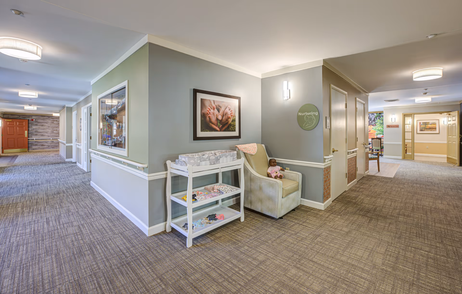 A well-lit hallway in a senior living facility with carpeted floors and light gray walls. On the right side, there is a cushioned armchair with a doll sitting on it and a white shelving unit holding various items. Above the chair is a framed picture of adult hands holding baby feet and a round sign on the wall that reads 'Nurturing.' The hallway extends with multiple doors and ceiling lights, and a red door is visible at the far end.
