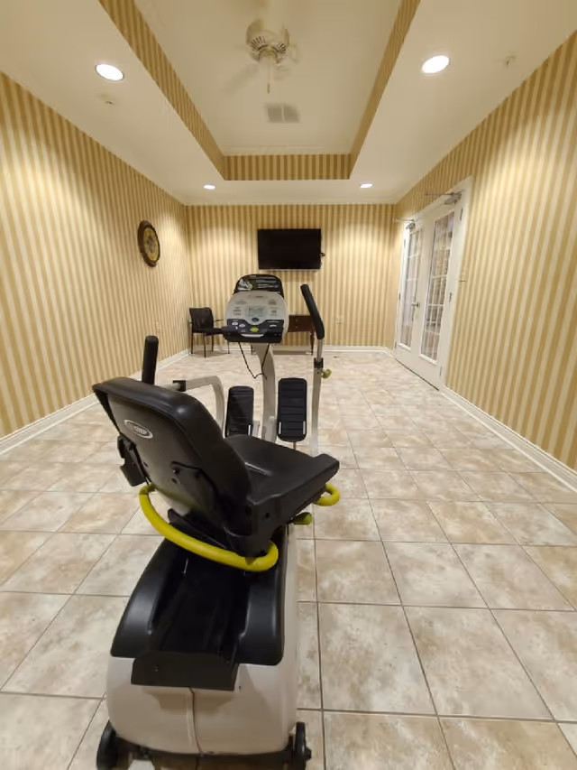 A small exercise room with beige and white striped wallpaper and tiled floor. The room contains a recumbent exercise bike facing a wall-mounted flat screen TV. There is a black chair and a clock on the wall to the left. The ceiling has recessed lighting and a ceiling fan. Double glass doors are visible on the right side of the room.
