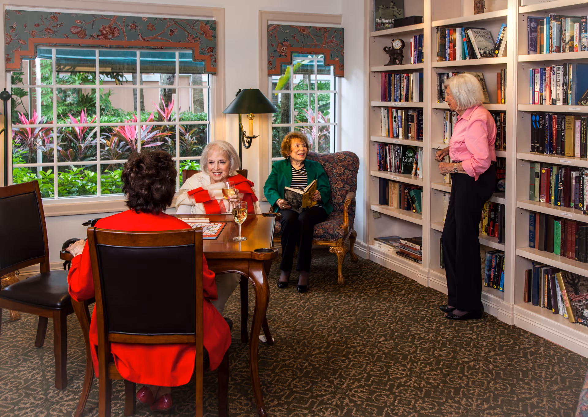 A cozy library room with four elderly women. Two women are seated at a wooden table with wine glasses, one woman is sitting in an armchair reading a book, and another woman is standing near a bookshelf filled with books. Large windows show greenery outside, and the room has patterned carpet and floral window valances.