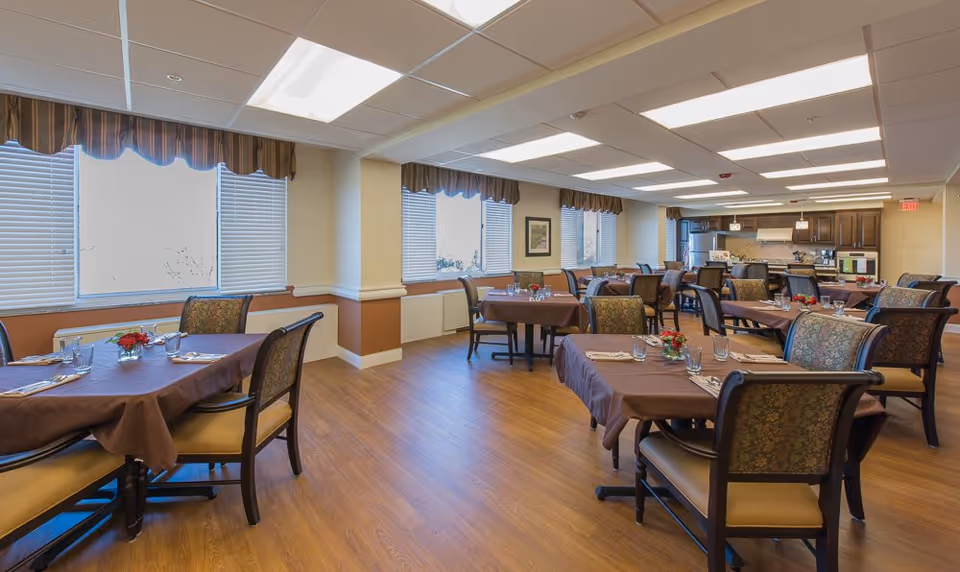 A spacious dining room with multiple tables covered in brown tablecloths, each set with glasses, napkins, and small floral centerpieces. The room has large windows with blinds and valances, wood flooring, and a kitchen area with dark wood cabinets and stainless steel appliances in the background.