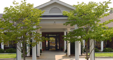Entrance to a building with a covered walkway supported by white columns and two green leafy trees on either side.
