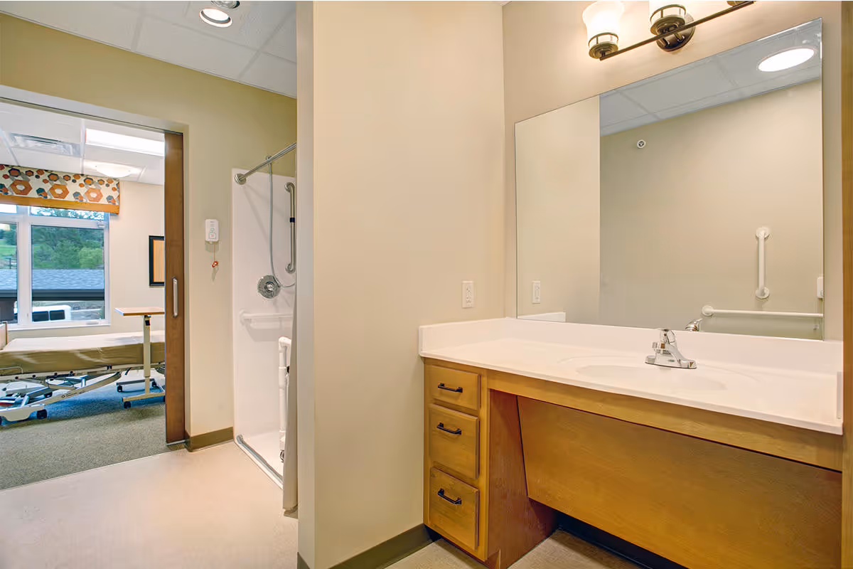 Accessible bathroom with a wooden vanity, sink and large mirror, a roll-in shower with grab bars, and a view into an adjacent room with a hospital-style bed.