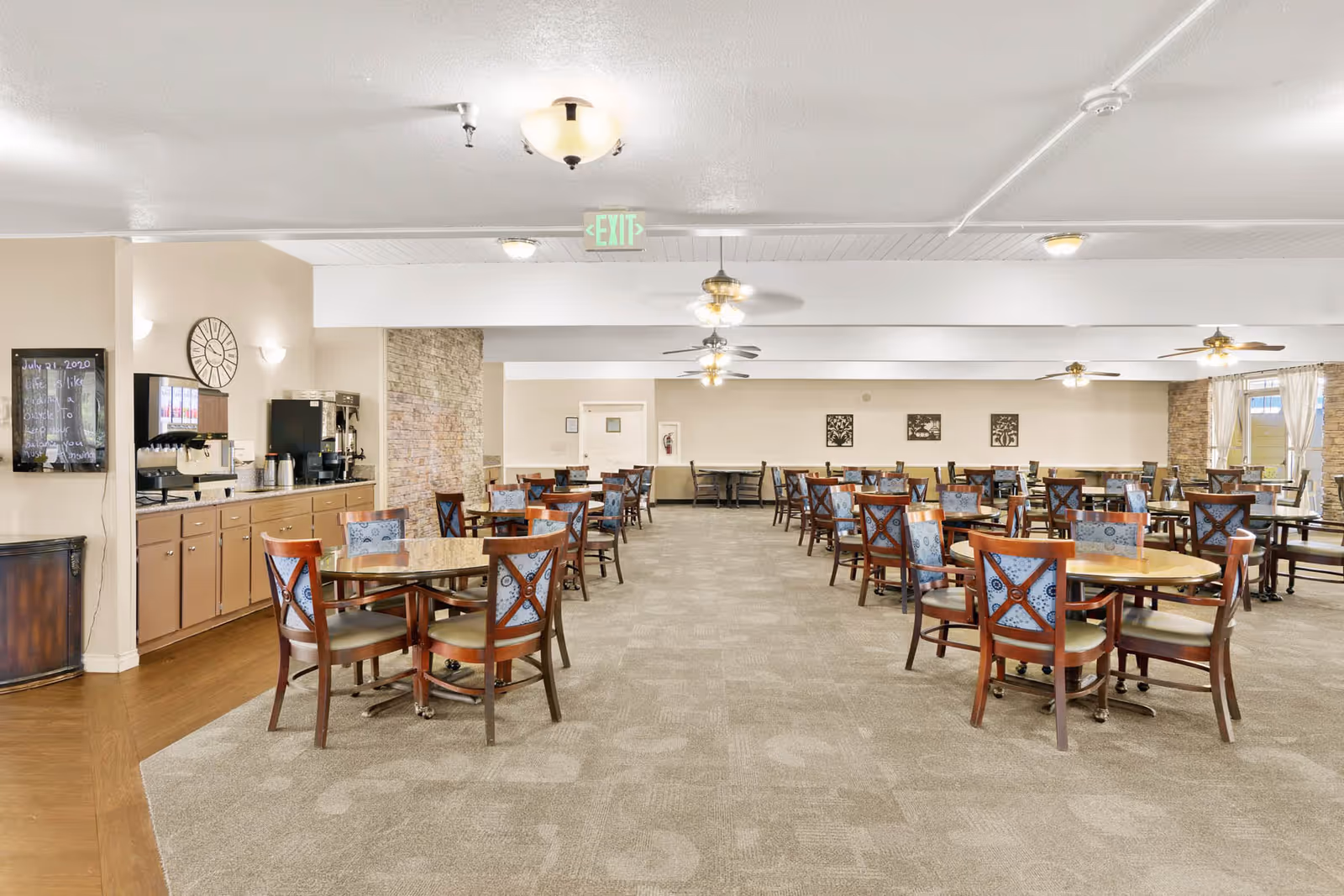 A spacious dining room in a senior living facility with multiple wooden tables and chairs arranged neatly. The room features beige walls, carpeted flooring, ceiling fans with lights, and large windows with curtains allowing natural light. On the left side, there is a beverage station with coffee machines and a soda dispenser. A clock and a framed board with writing are mounted on the wall near the beverage station.