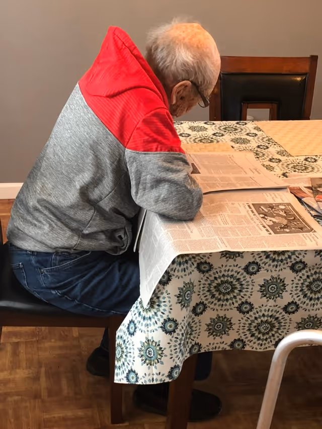 An elderly man with gray hair and glasses is sitting at a dining table covered with a patterned tablecloth, reading a newspaper. He is wearing a gray and red jacket and blue jeans. The room has wooden flooring and a gray wall in the background.