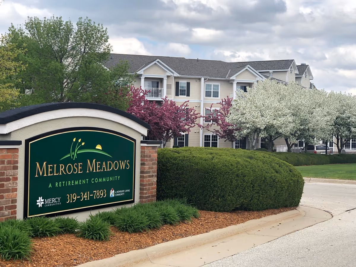 Entrance sign reading 'Melrose Meadows' with landscaped shrubs and the retirement community building in the background.