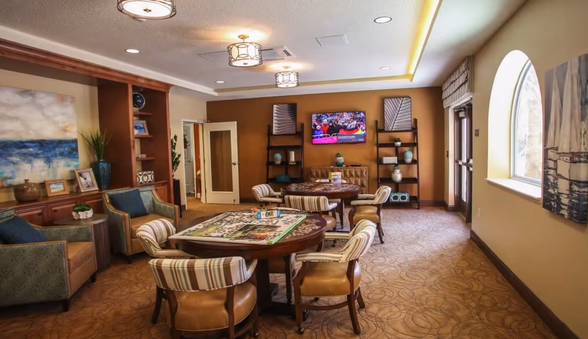 A cozy common area with two round tables surrounded by cushioned chairs. The room features warm brown walls, patterned carpet, and decorative shelving with vases and plants. A flat-screen TV is mounted on the far wall, and large windows let in natural light.