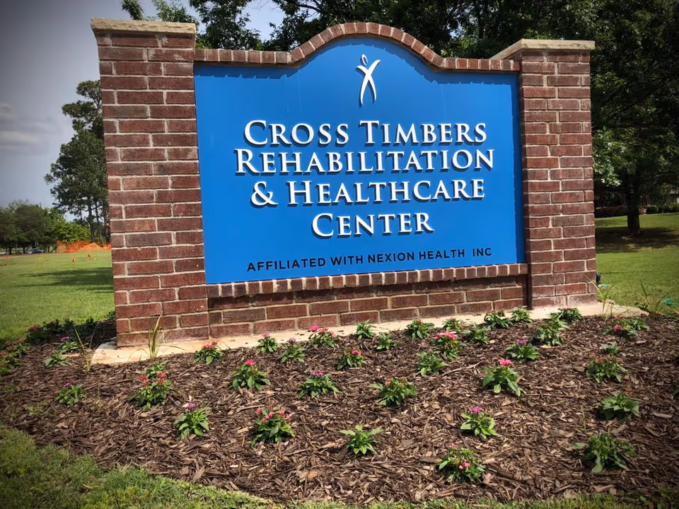 Brick-framed blue entrance sign that reads "Cross Timbers Rehabilitation & Healthcare Center" set in a landscaped lawn with flowers.