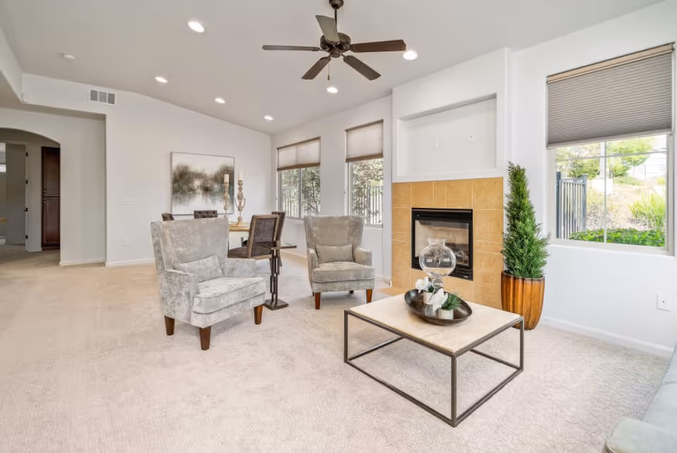 A bright and spacious living room with beige carpet, two gray upholstered armchairs, a rectangular coffee table with decorative items, a fireplace with a tiled surround, large windows with blinds, a potted plant, and a ceiling fan with lights.
