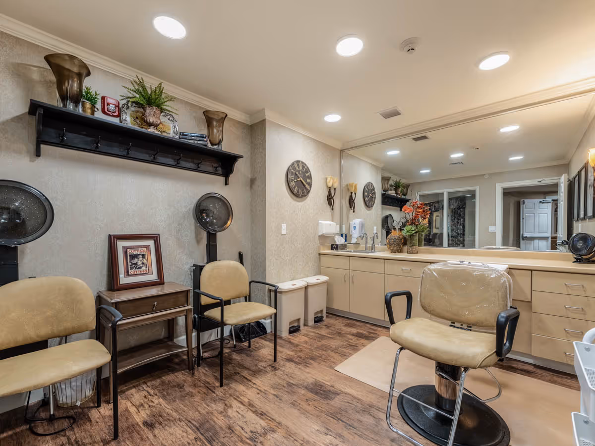 Interior of a salon room with a salon chair in the foreground, two beige chairs against the wall, two hair dryers, a small wooden table, a large mirror above a counter with cabinets, decorative plants, and wall clocks.