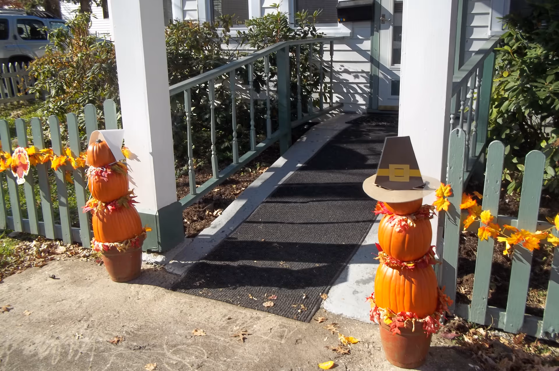 Entrance to a building with a ramp and green picket fence decorated with autumn leaves garlands. Two stacked pumpkin decorations with fall leaves and one topped with a pilgrim hat flank the ramp.