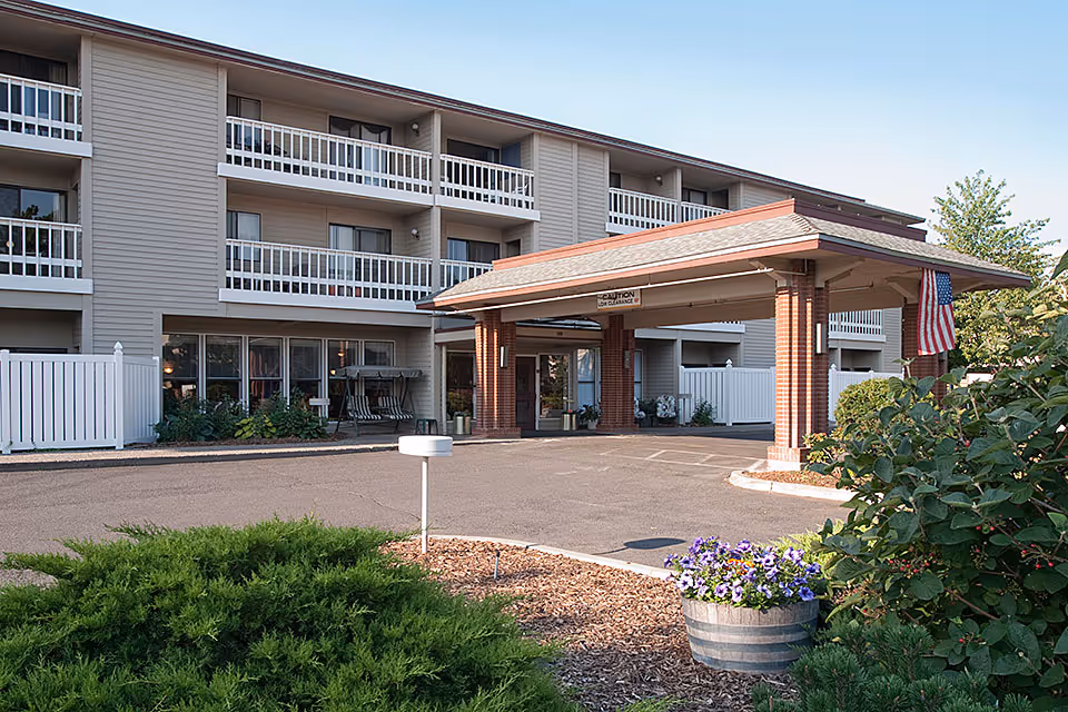 Exterior view of a multi-story senior living facility with balconies, a covered entrance supported by brick columns, an American flag, and landscaped greenery including bushes and a flower pot with purple flowers.