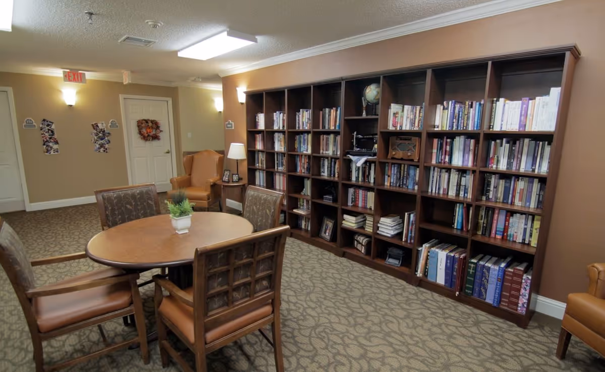 A cozy reading room with a large wooden bookshelf filled with books along one wall. In front of the bookshelf is a round wooden table with four cushioned chairs around it. There is a small potted plant on the table. The room has beige walls, patterned carpet, and soft lighting from wall sconces and a table lamp next to a comfortable armchair. Two white doors and an exit sign are visible in the background.