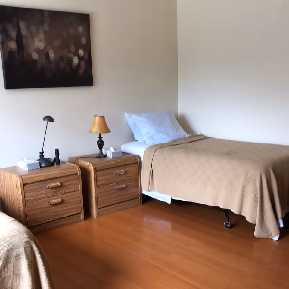 A simple bedroom with a single bed covered with a beige blanket and a white pillow. Next to the bed are two wooden nightstands, one with a small lamp and the other with a desk lamp and some tissues. A framed picture with blurred lights hangs on the wall above the nightstands. The floor is wooden.