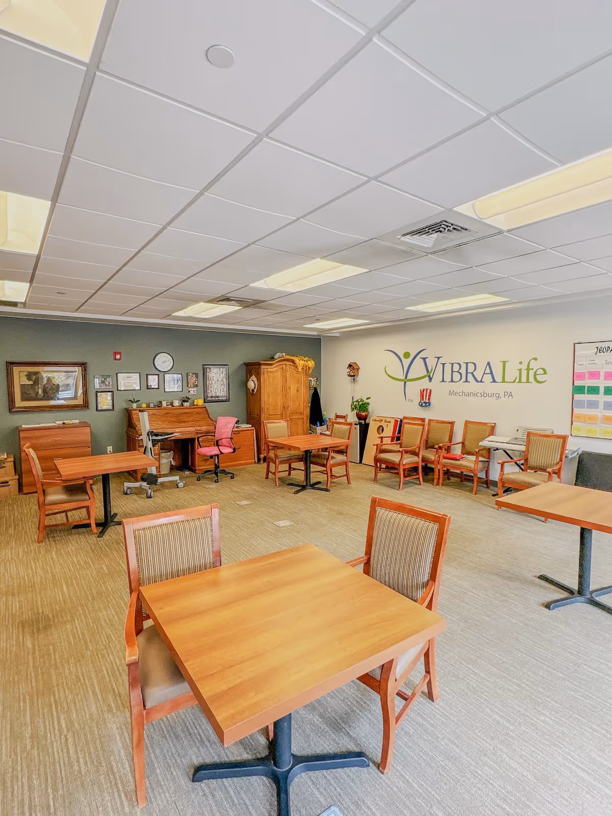 A well-lit room with multiple wooden tables and chairs arranged for seating. The walls have decorations including framed pictures, a clock, and a large VibraLife logo with the text 'Mechanicsburg, PA'. There is a wooden cabinet, a desk with a pink office chair, and a keyboard in the corner. The room has a carpeted floor and a drop ceiling with fluorescent lights.