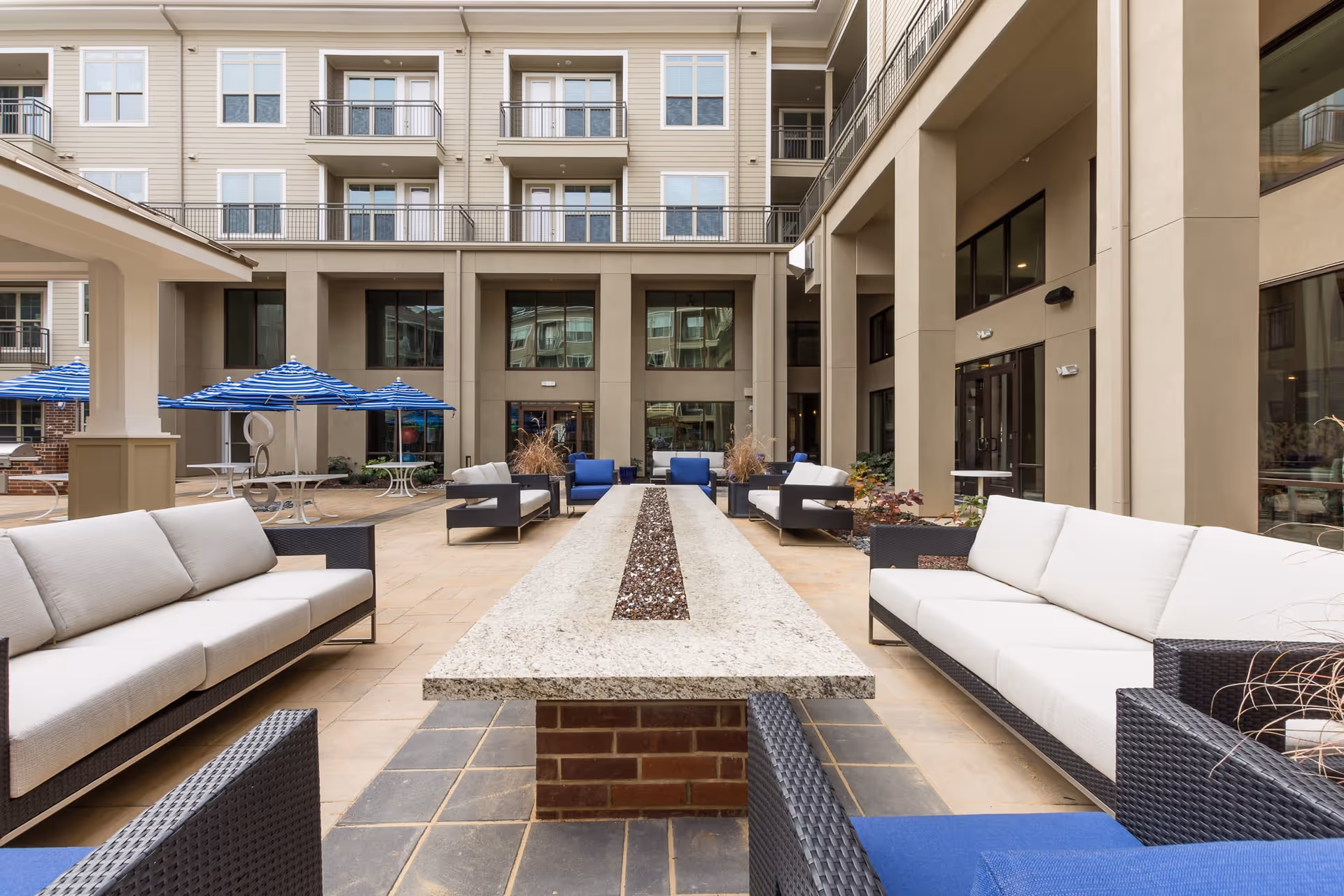 Outdoor courtyard area of an apartment complex with cushioned wicker sofas and chairs arranged around a long rectangular stone fire pit table. Blue and white striped umbrellas shade round tables in the background, with beige multi-story apartment buildings surrounding the courtyard.
