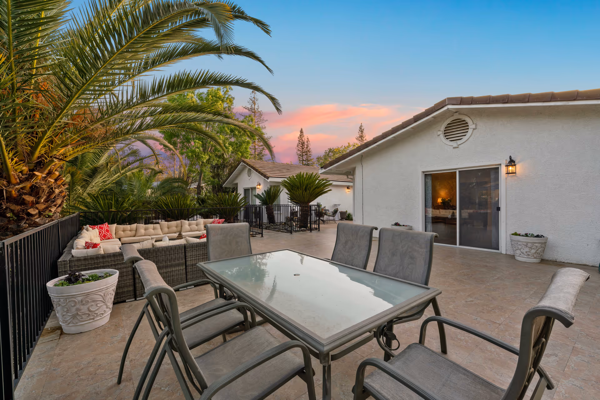 Outdoor patio area at a senior living facility with a glass-top dining table and six chairs in the foreground, a large cushioned sectional sofa with red and white pillows along a black metal railing, potted plants, palm trees, and a white building with sliding glass doors and exterior lights at sunset.