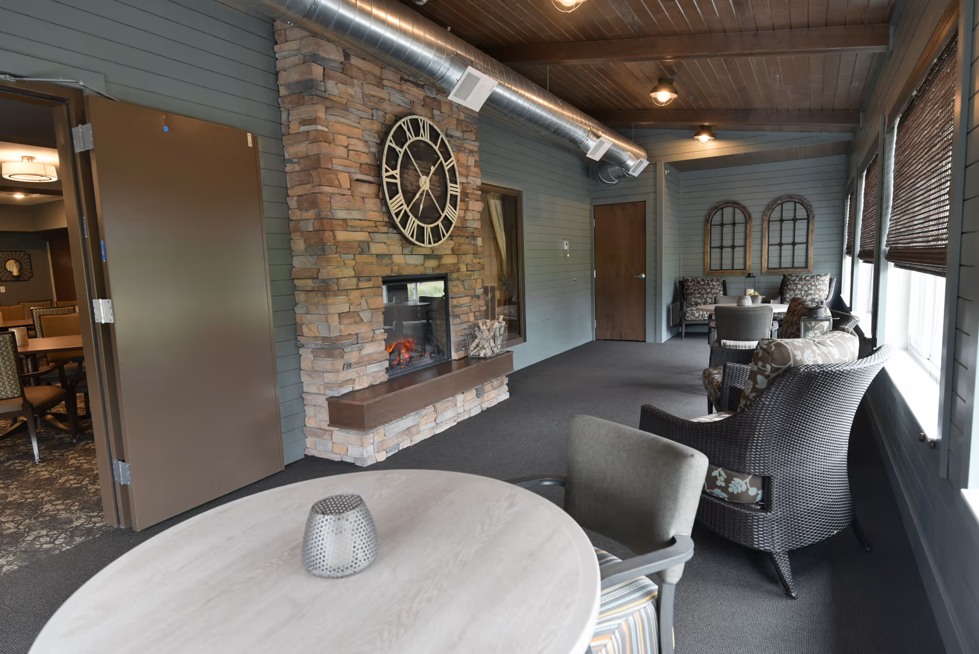 A cozy indoor seating area with wicker chairs and patterned cushions arranged around small tables. There is a stone fireplace with a large clock above it on the wall. The room has wooden ceiling beams, a visible metal air duct, and windows with woven blinds letting in natural light. A door leads to another room with dining tables and chairs.
