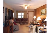 Cozy living room with a ceiling fan, floral patterned sofa, armchair, wooden side tables with lamps, a large TV on a wooden stand, and a dining table with chairs in the foreground. The walls are wood-paneled and there is a window with curtains and a door in the background.