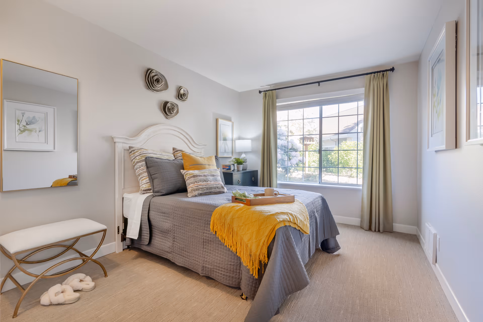 Bright, neatly styled bedroom with a bed dressed in gray and yellow linens, a large window with curtains, and decorative wall art.