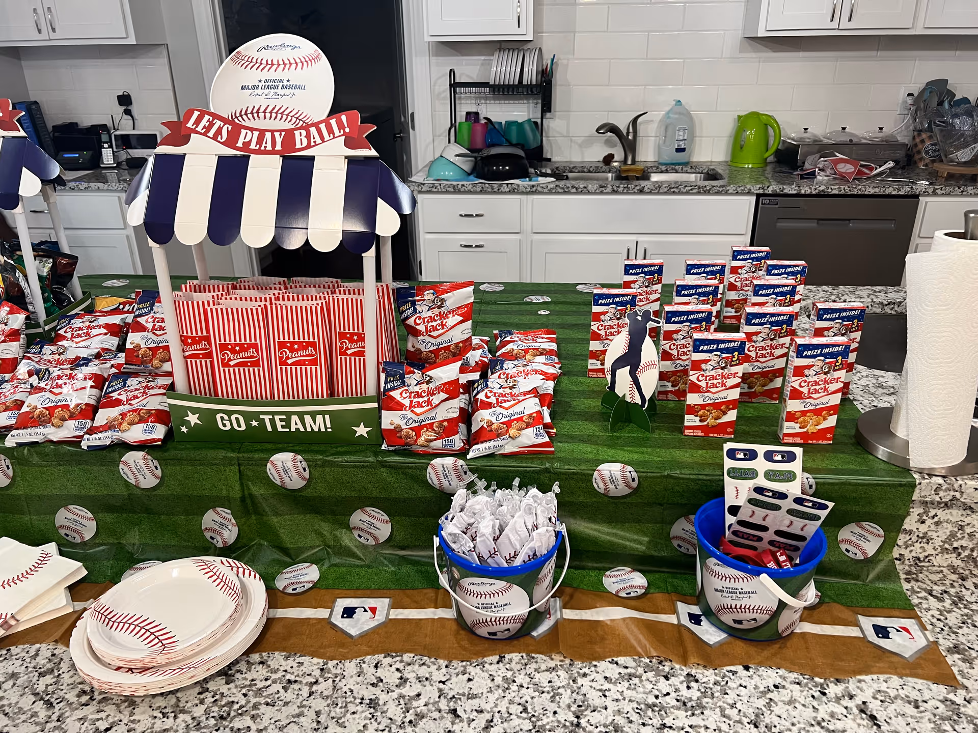 Baseball-themed snack display on a kitchen counter with Cracker Jack bags, striped popcorn boxes, baseball plates and themed decorations.