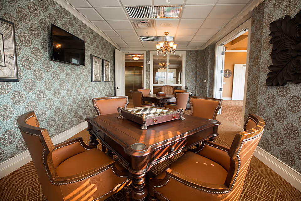 A cozy interior room with a wooden table and four brown leather chairs arranged around it. On the table is a chess/checkers board. The room has patterned wallpaper, a wall-mounted TV, framed pictures, and a chandelier hanging from the ceiling. There is an open doorway leading to another room with carpeted floors.