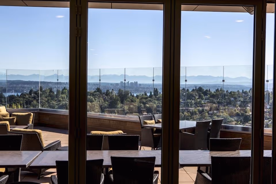 View through glass doors of an outdoor patio area with tables and chairs, overlooking a scenic landscape of trees, a distant city skyline, and mountains under a clear blue sky.