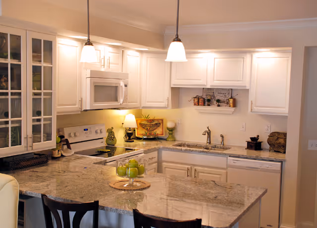 A modern kitchen with white cabinets, granite countertops, and stainless steel appliances including a microwave and stove. There are two pendant lights hanging over a kitchen island with a glass bowl of green apples. The kitchen also features a double sink, a dishwasher, and decorative items on the countertops and shelves.