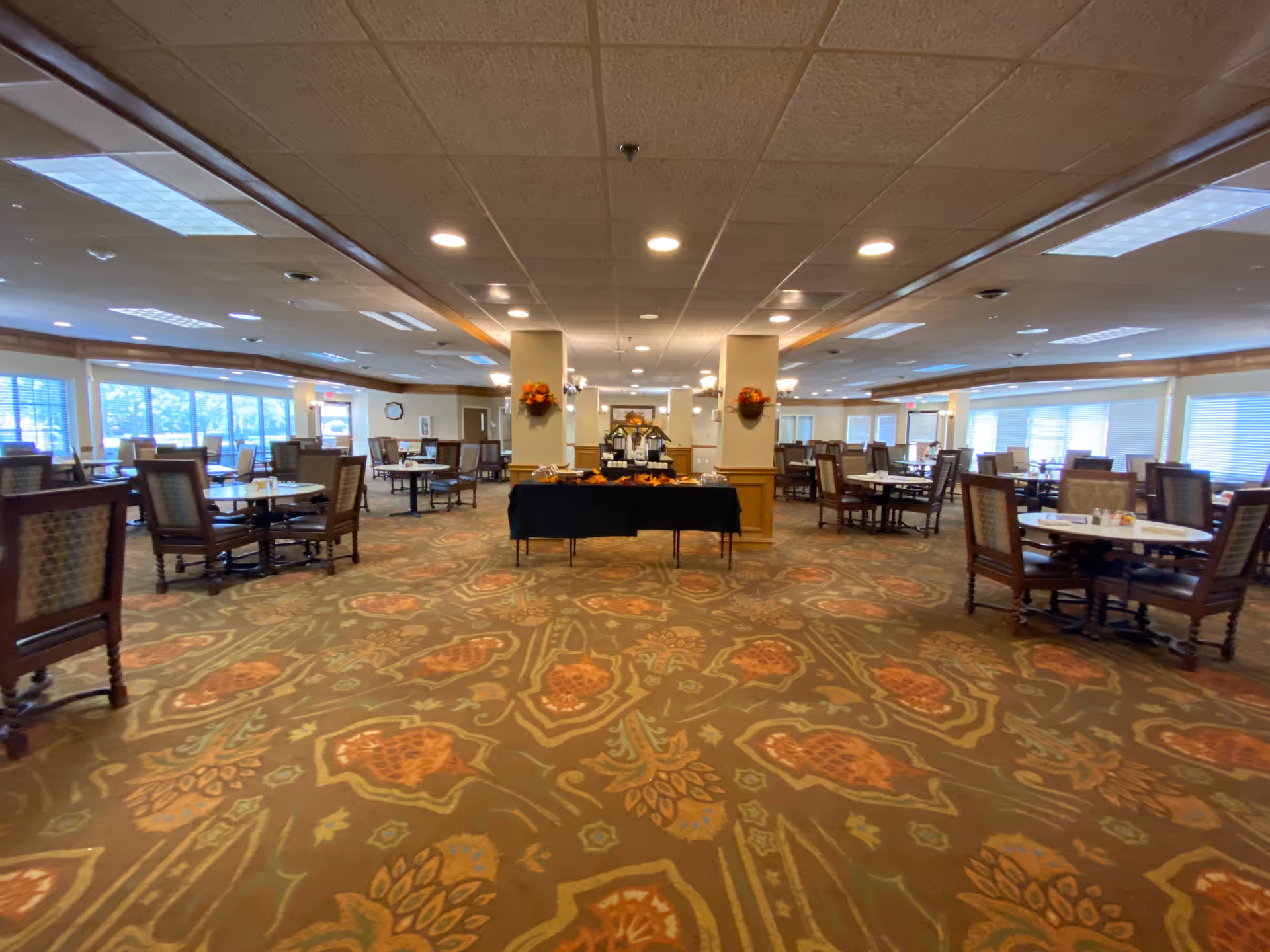 A spacious dining room with multiple round tables and cushioned chairs arranged neatly. The room has large windows allowing natural light to fill the space, and a central table covered with a black cloth displaying various items. The carpet has a floral pattern in warm tones, and the ceiling has recessed lighting.