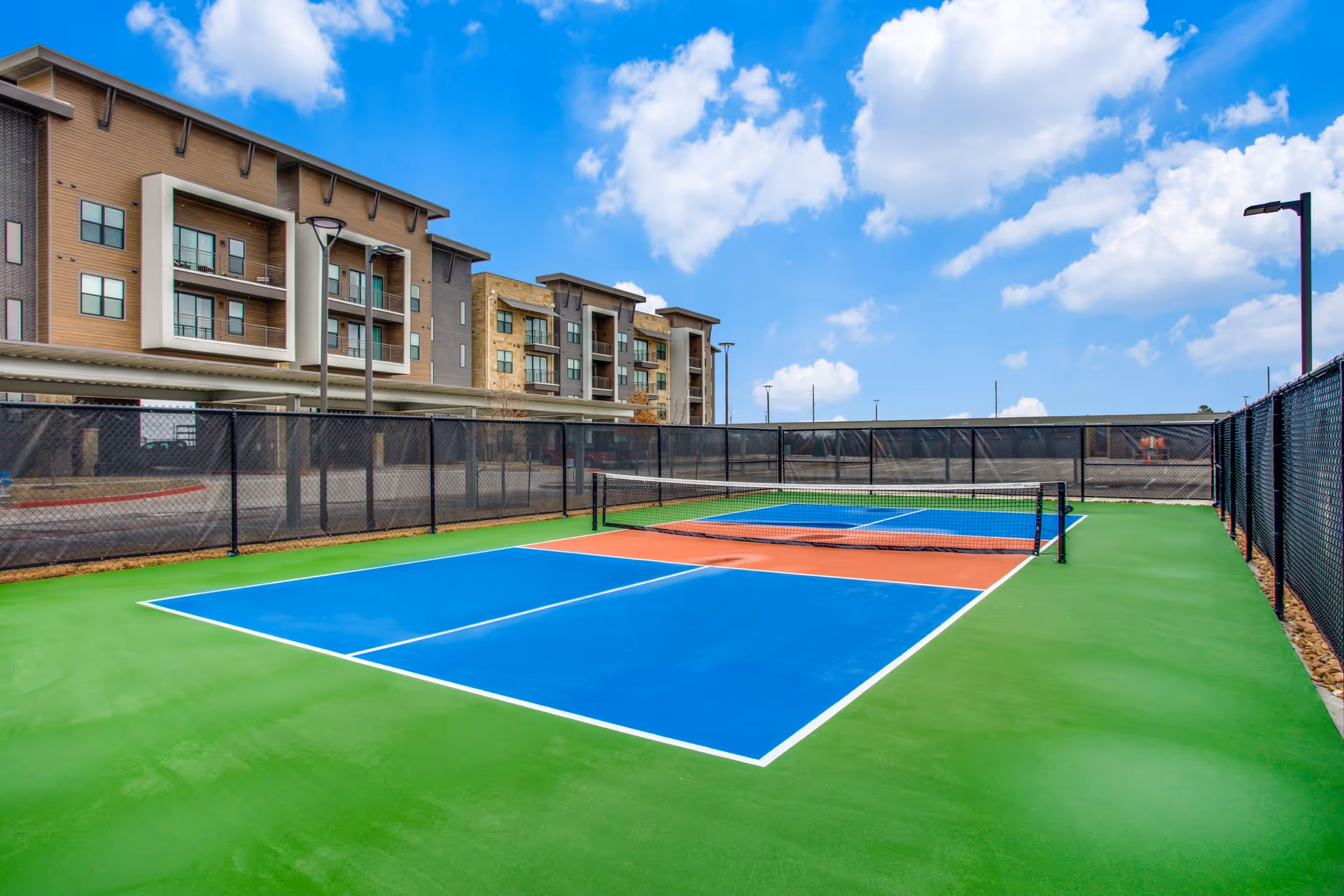 Colorful outdoor tennis/pickleball court with blue and orange playing surfaces, fenced and set in front of a multi-story apartment building under a blue sky.