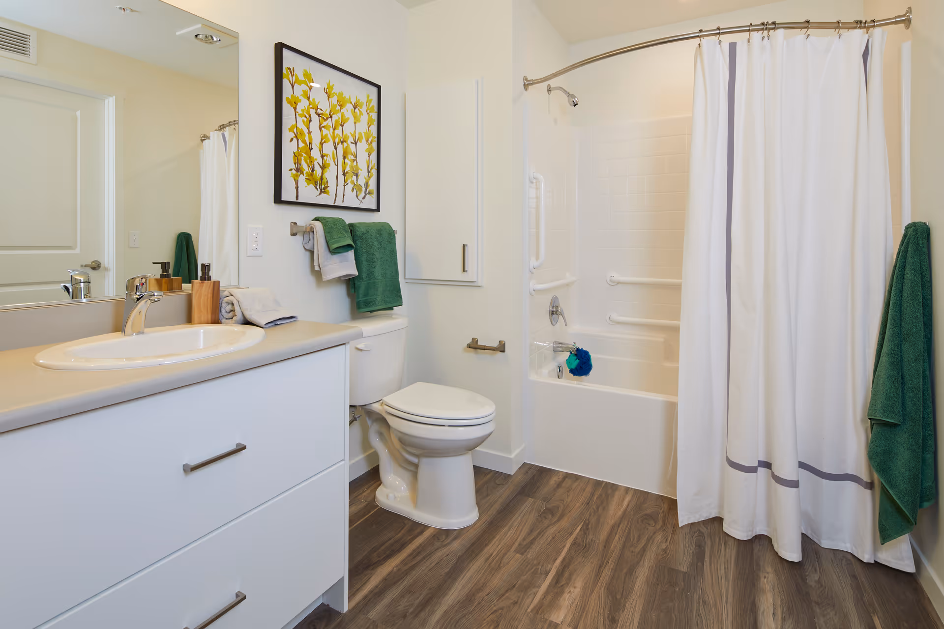 A clean and modern bathroom featuring a white vanity with a sink, a large mirror, a toilet, and a bathtub with a white shower curtain. The bathroom has wood-look flooring, white walls, and green towels hanging on racks. There is a framed artwork of yellow flowers on the wall above the toilet.