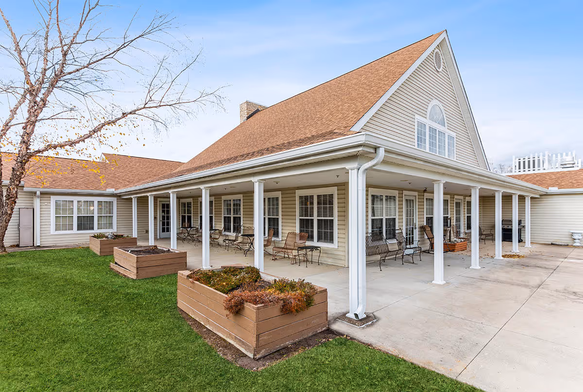 Exterior view of a single-story building with a covered porch supported by white columns. The porch has several metal chairs and benches arranged along the wall. There are three large wooden planter boxes with some plants in front of the porch. The building has beige siding and a brown shingled roof. A leafless tree is visible on the left side, and the sky is clear with a few clouds.