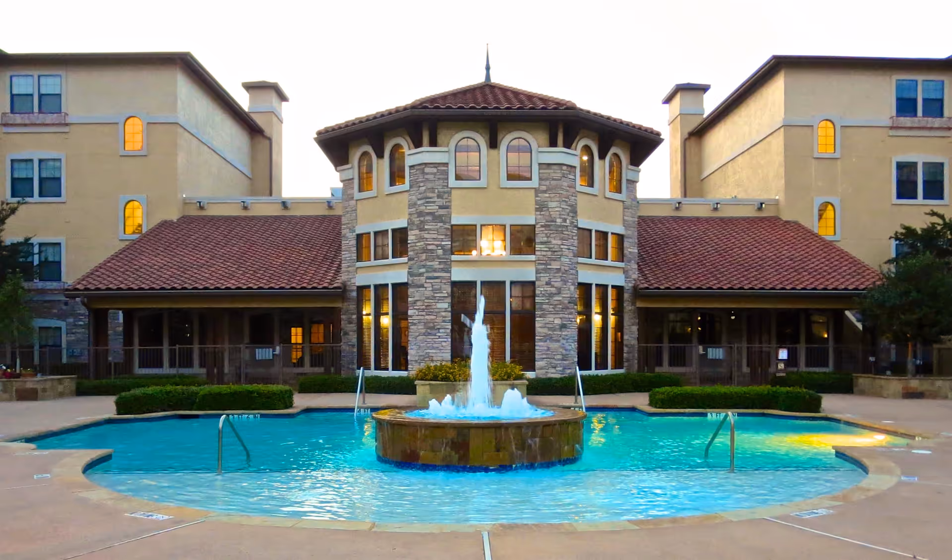 Front view of a Mediterranean-style residential building with a central stone tower and a fountain-filled swimming pool in the foreground.