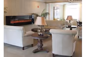 Bright lounge with upholstered armchairs and sofas arranged around wooden side tables and lamps, with a wall-mounted electric fireplace.