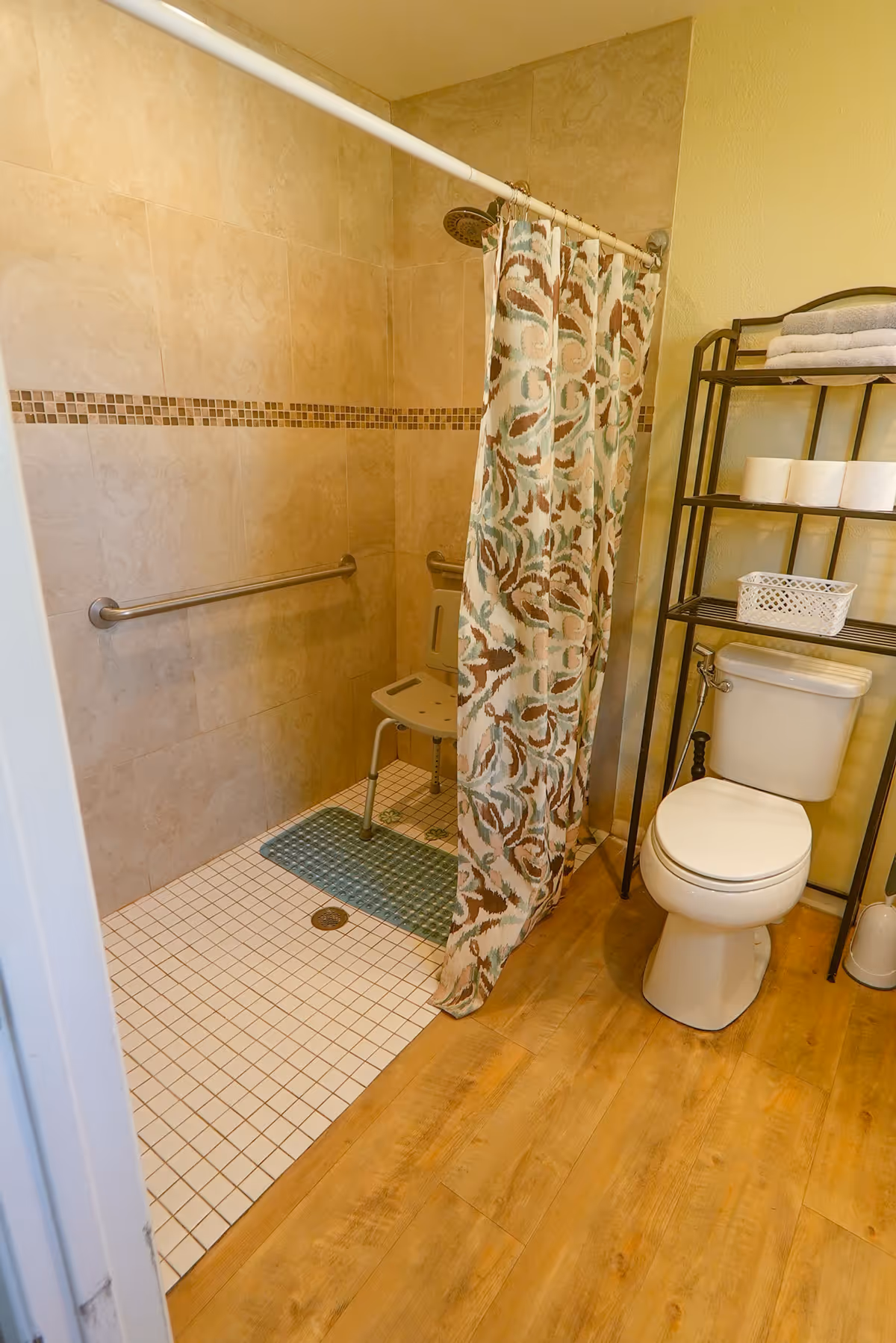 A bathroom with a walk-in tiled shower featuring a shower chair and grab bars, a patterned shower curtain, a toilet next to a black metal shelving unit holding towels, toilet paper, and a basket, and wood-look flooring.