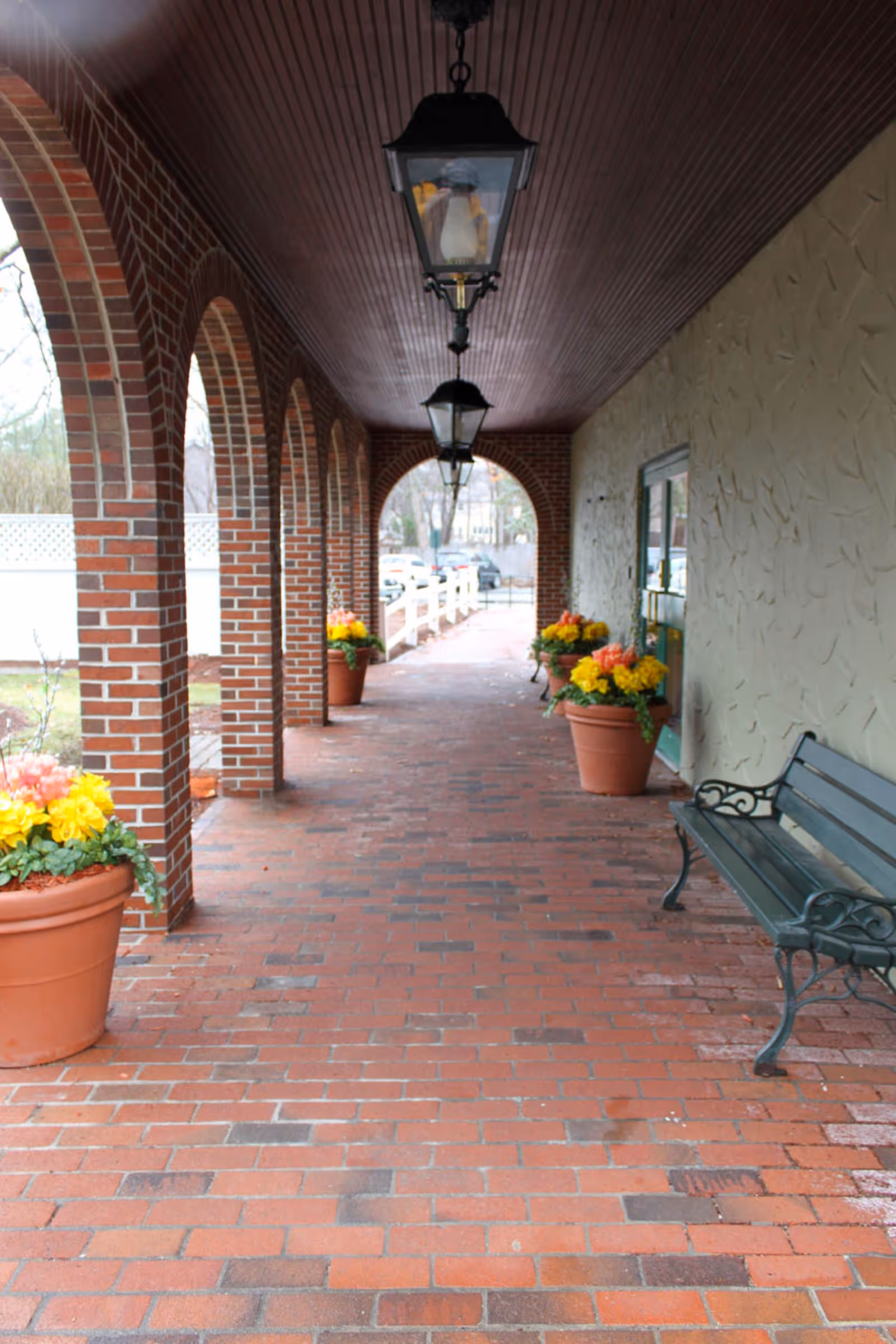 Covered brick walkway with arched brick columns on the left and a textured wall on the right. There are hanging lantern-style lights on the wooden ceiling, large flower pots with colorful flowers along the walkway, and a black metal bench on the right side.