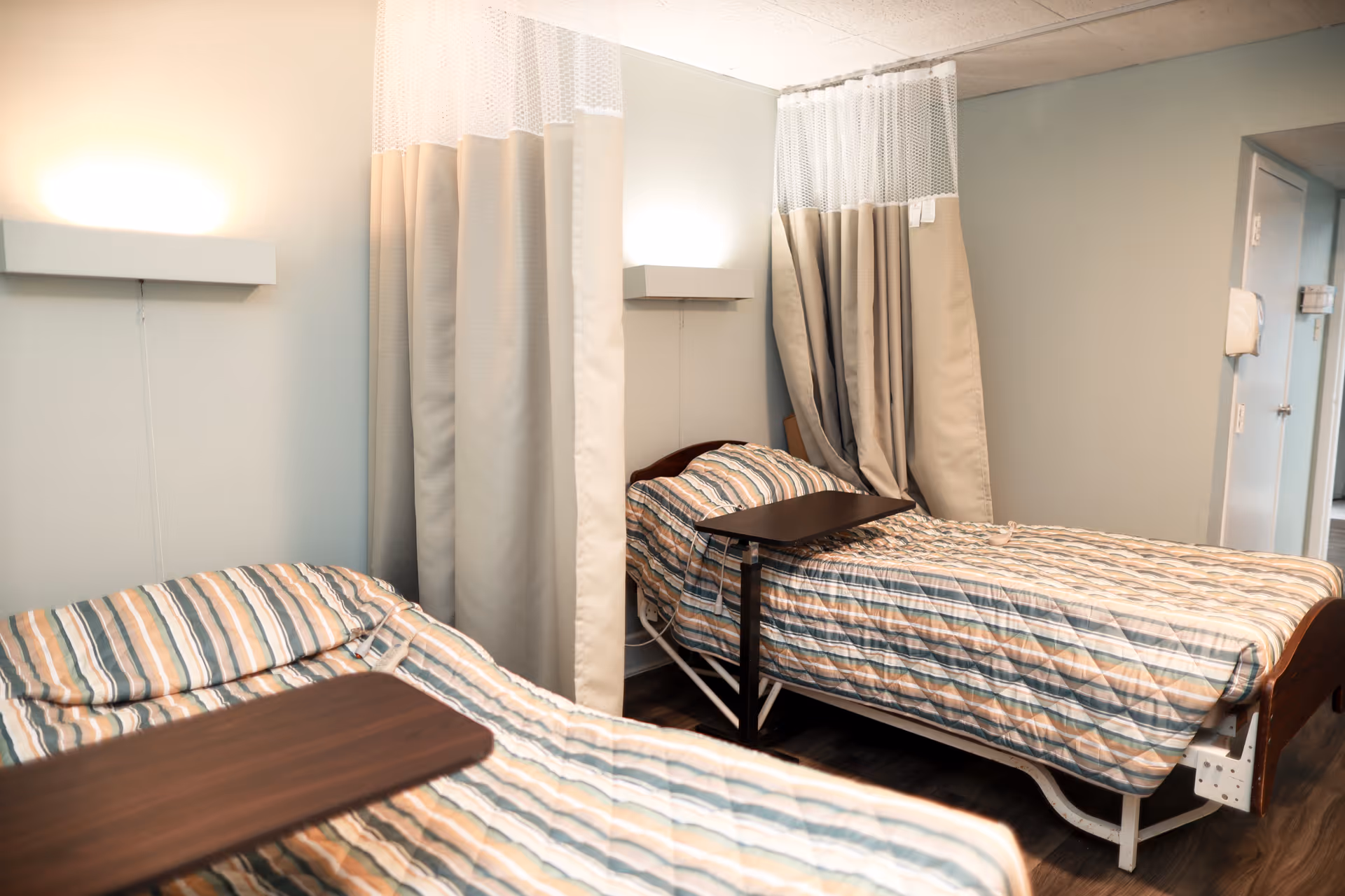 A room with two hospital-style beds separated by privacy curtains. Each bed has a striped bedspread and an overbed table. The walls are light-colored and there are wall-mounted lights above each bed.