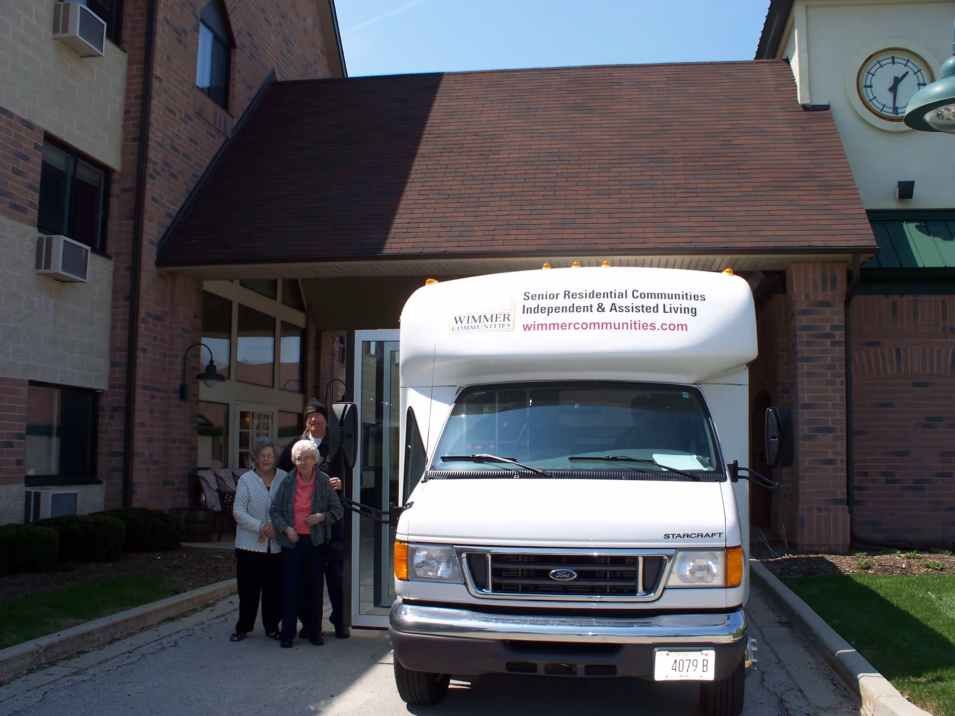 A white shuttle bus parked in front of a senior living facility entrance with three elderly people standing beside it. The bus has signage for Wimmer Communities, advertising senior residential communities for independent and assisted living. The building has brick and stone exterior walls with windows and a clock tower visible on the right side.
