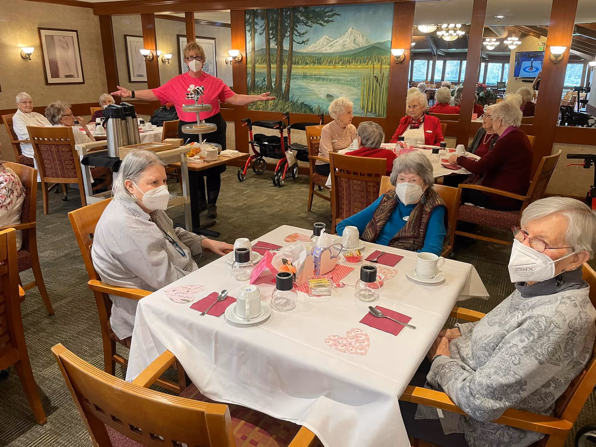 Elderly residents wearing masks seated around a decorated dining table while a staff member stands nearby in a dining room with a large landscape mural.