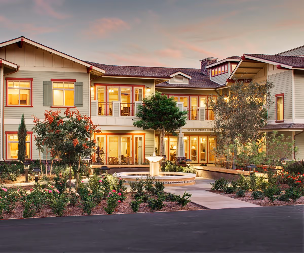 Exterior view of Santa Teresita Assisted Living Cottages at dusk, showing a two-story building with warm interior lights, a central fountain, landscaped garden with trees and flowers, and a paved walkway leading to the entrance.