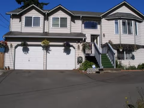 Front exterior view of a two-story residential building with a double garage, multiple windows, and a staircase leading to the main entrance. The house is light-colored with dark roofing and has hanging flower baskets and some greenery around the entrance.