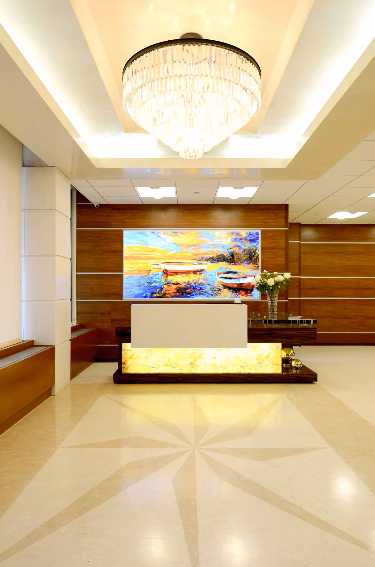 A modern reception area with a large chandelier hanging from the ceiling, a backlit reception desk with a glowing front panel, a colorful painting of boats on water mounted on a wooden wall behind the desk, and a vase of white flowers on the desk.