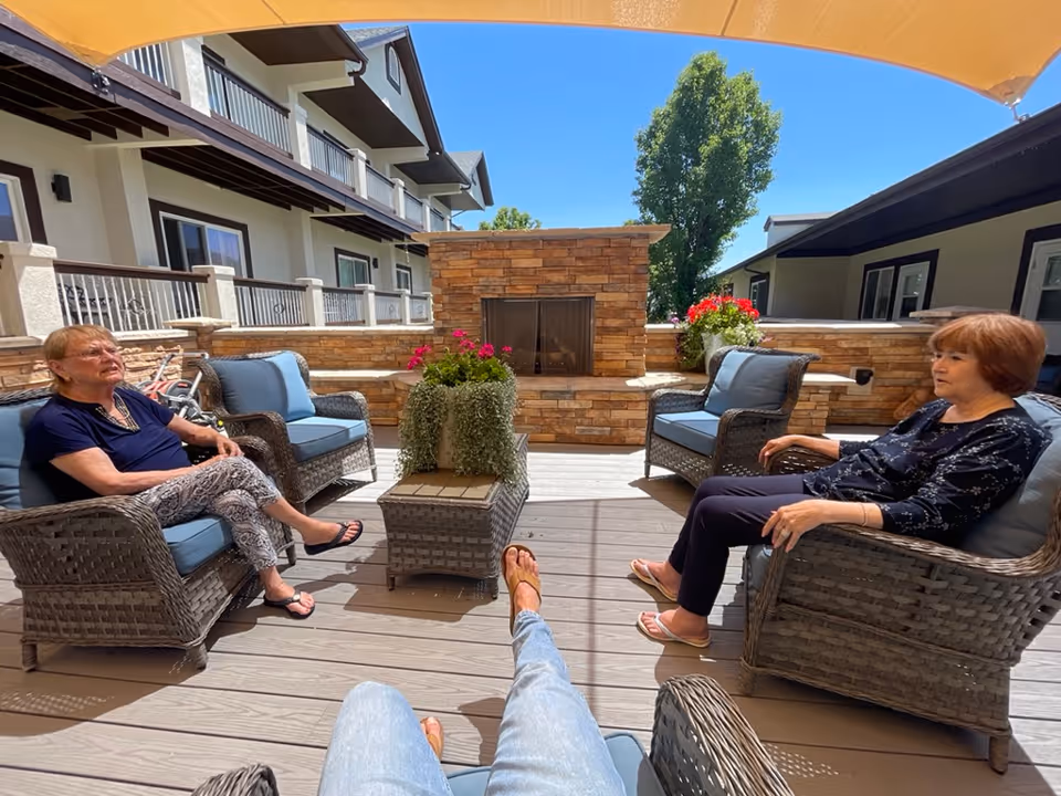 Two elderly women sitting on cushioned wicker chairs on a patio with a stone fireplace and potted plants, under a yellow canopy. The patio is part of a multi-story building with balconies and railings, and there is a clear blue sky in the background.